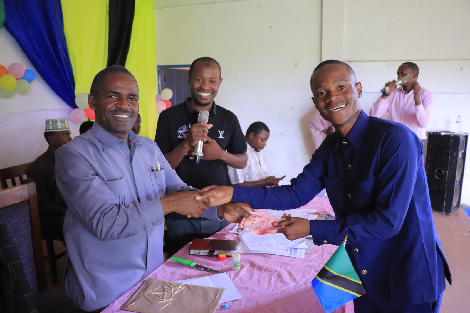 The Regional Education Officer is handing over cash to the Coordinator of the "No Child Left Behind" campaign along with the principal supervisor of training for graduating primary school students.